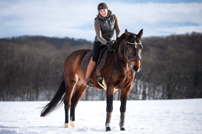 Eine Frau sitzt auf ihrem braunen Pferd in einer Schneelandschaft.