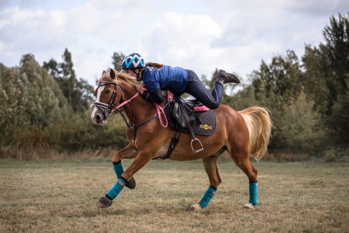 Eine Frau springt auf ihr galoppierendes Pony auf. Das Pony ist fuchsfarbend mit heller Mähne und Schweif.