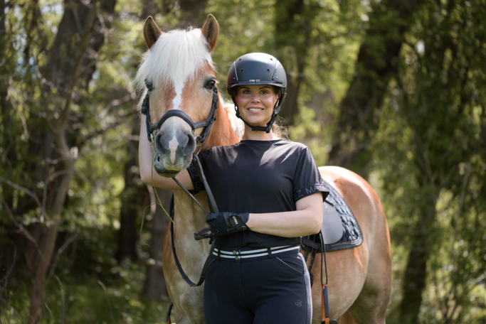 Eine lächelnde Frau steht neben einem Pferd, einem Haflinger. Beide schauen in die Kamera. Die Frau hat ihre Hand am Kopf des Pferdes.
