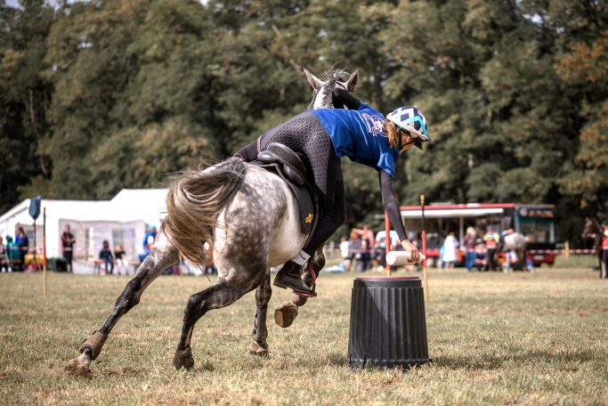 Frau galoppiert mit Pferd an einer Tonne vorbei und nimmt dabei eine Flasche von der Tonne auf. Das Pferd ist ein Schimmel.